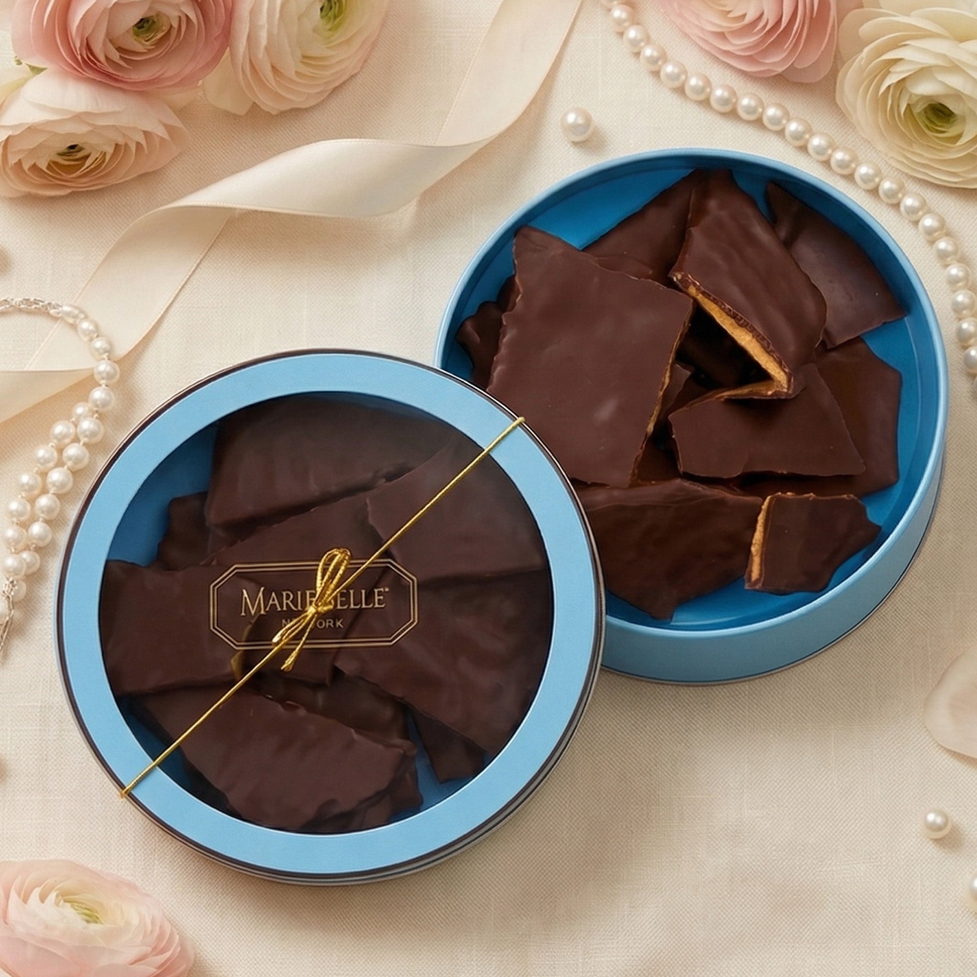 Open blue tin filled with chocolate pieces on a decorative surface with flowers and pearls.