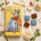 Chocolate box with illustrations of a woman and chocolates on a decorative surface with flowers and pearls.